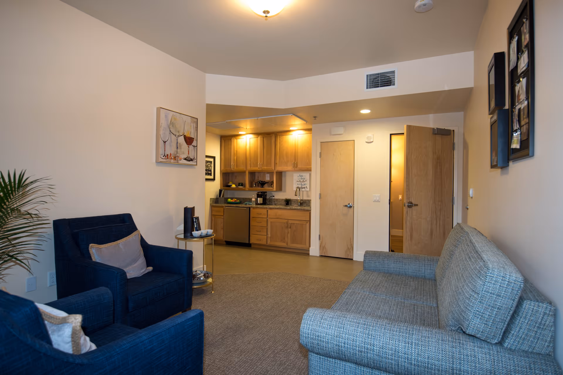 A cozy living area in a senior living facility with two blue armchairs and a gray sofa arranged around a small round side table. The room features light-colored walls with framed artwork, a potted plant, and an open view into a small kitchen area with wooden cabinets, a dishwasher, and a sink. Two wooden doors are visible, one slightly open, revealing a glimpse of another room.