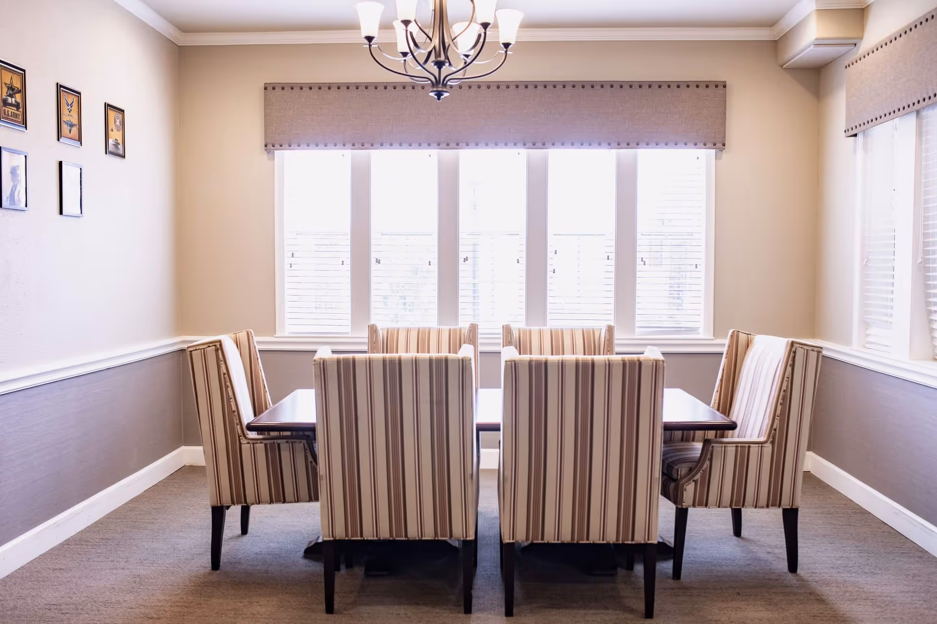 A bright dining room with a rectangular wooden table surrounded by six striped upholstered chairs. The room has beige walls with white trim, a chandelier hanging from the ceiling, and large windows with blinds letting in natural light. Several framed pictures hang on one wall.