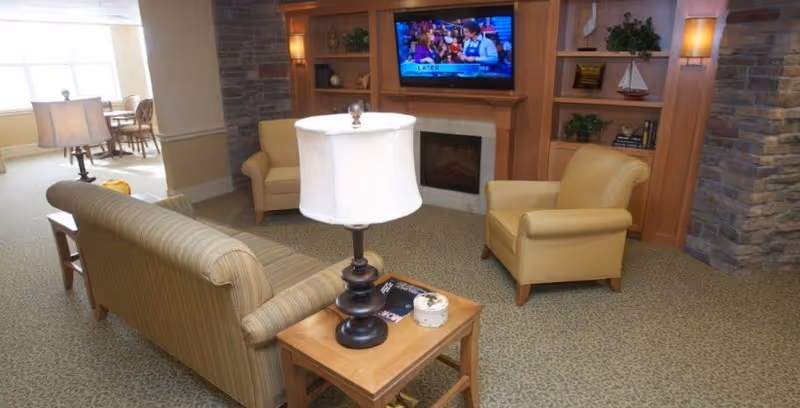 A cozy living room area with a striped sofa, two beige armchairs, a wooden side table with a lamp, and a built-in wooden entertainment center with a TV mounted above a fireplace. The room has carpeted flooring and stone accents on the walls.