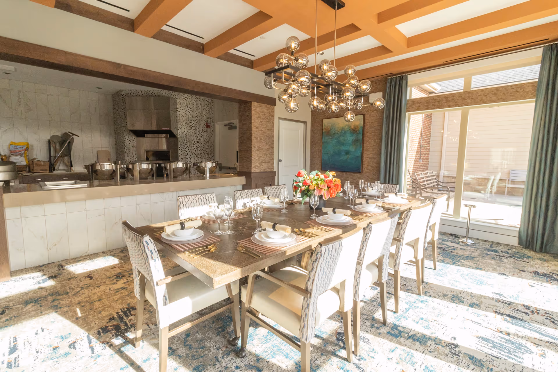 A bright dining room with a large wooden table set for ten people with plates, glasses, napkins, and cutlery. A modern chandelier with multiple glass bulbs hangs above the table. The room has large windows with curtains allowing natural light to fill the space. In the background, there is a kitchen area with stainless steel appliances and a tiled backsplash.