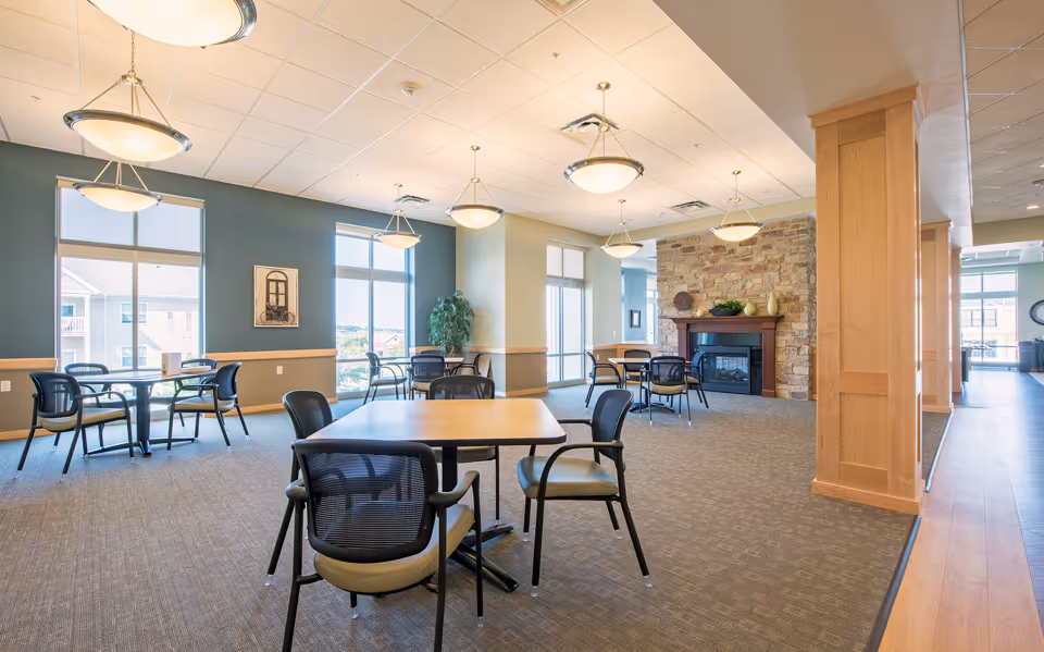 A spacious and well-lit common dining area with multiple square tables and black chairs arranged around them. The room features large windows allowing natural light to fill the space, a stone fireplace with decorative items on the mantel, and modern ceiling light fixtures. The walls are painted in soft blue and beige tones, and there is a wooden pillar and trim adding warmth to the room.