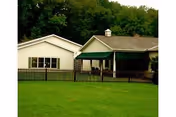 A single-story building with white siding and a green awning over a porch area, surrounded by a black picket fence and a large grassy lawn, with dense trees in the background.