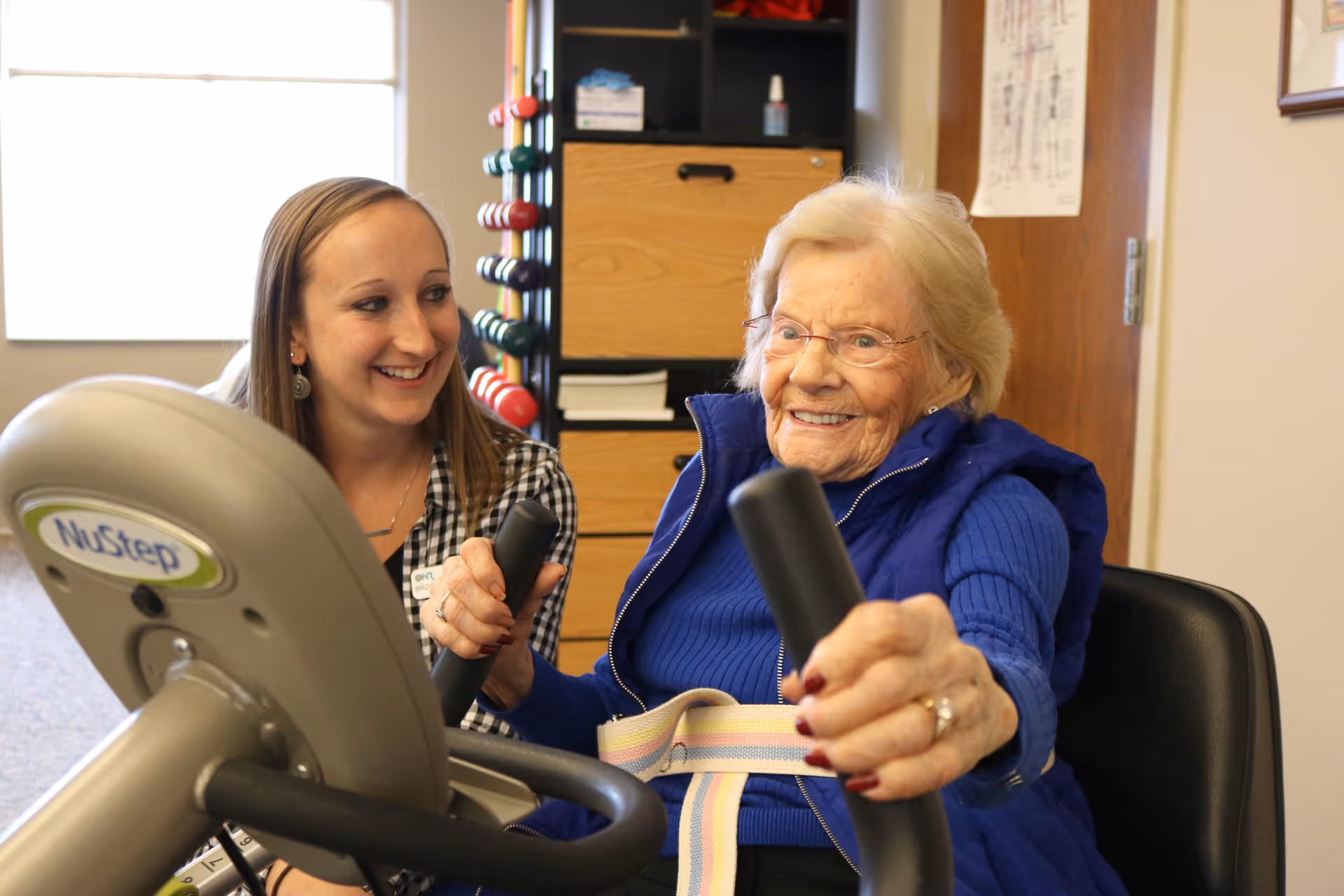 An elderly woman wearing a blue sweater and vest is exercising on a NuStep machine with a smiling female therapist assisting her in a therapy center room.
