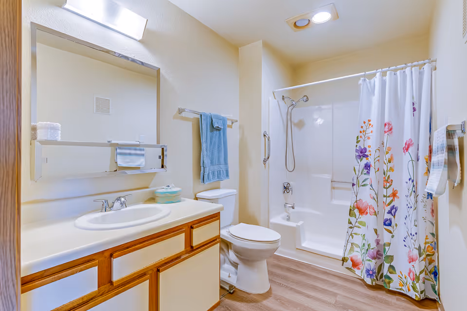 A clean and bright bathroom featuring a white sink with a wooden cabinet underneath, a large mirror with a light above it, a toilet, and a shower area with a floral-patterned shower curtain. A blue towel hangs on a rack above the toilet, and the floor has a wood-like finish.