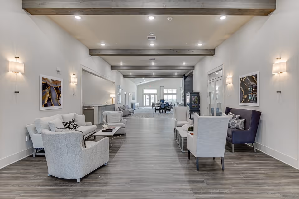 A spacious and modern living area in Auburn Assisted Living featuring comfortable seating arrangements with armchairs and sofas on either side of a wide walkway. The room has wooden ceiling beams, wall-mounted lights, abstract artwork, and a dining area visible in the background near large windows.