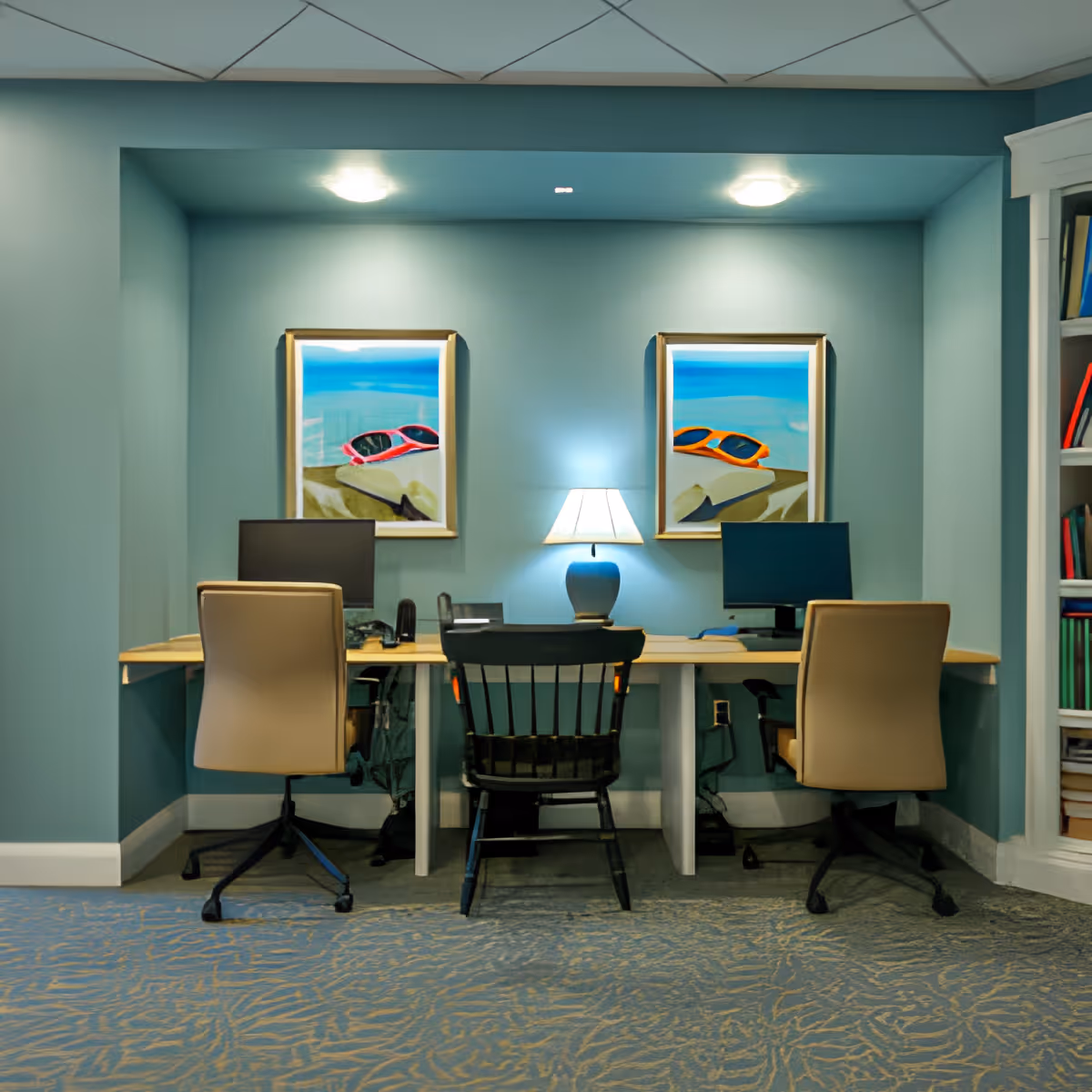 A small teal-painted interior workspace with two desks, three chairs, computer monitors and a lamp beneath two framed beach paintings.