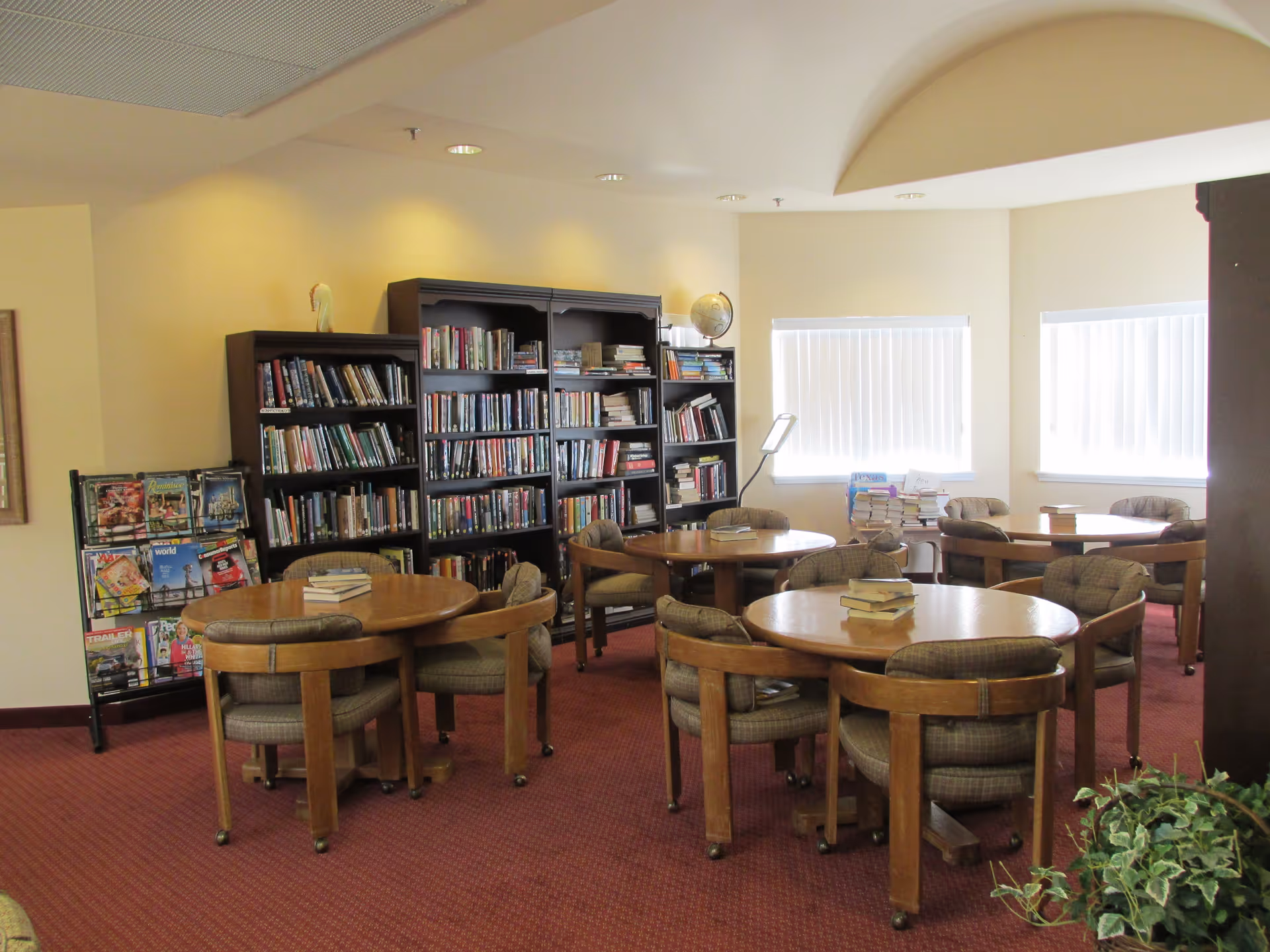 A cozy library or reading room with several round wooden tables surrounded by cushioned chairs. Bookshelves filled with books and a magazine rack are visible against the wall. Two windows with vertical blinds allow natural light into the room. A small plant is seen in the foreground.