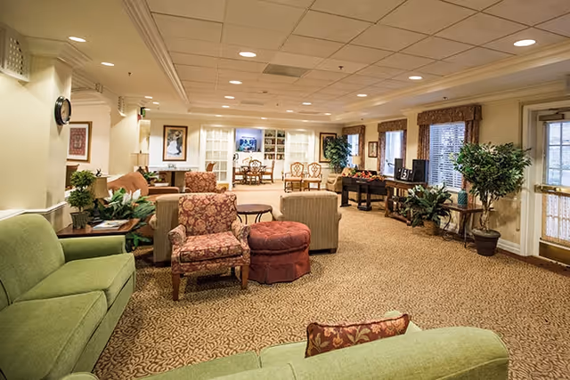 A spacious, well-lit living room area with patterned carpet, multiple upholstered chairs and sofas in green and floral prints, a round ottoman, side tables, potted plants, framed artwork on the walls, and large windows with blinds. In the background, there is a dining area with a table and chairs, and built-in shelves with decorative items.