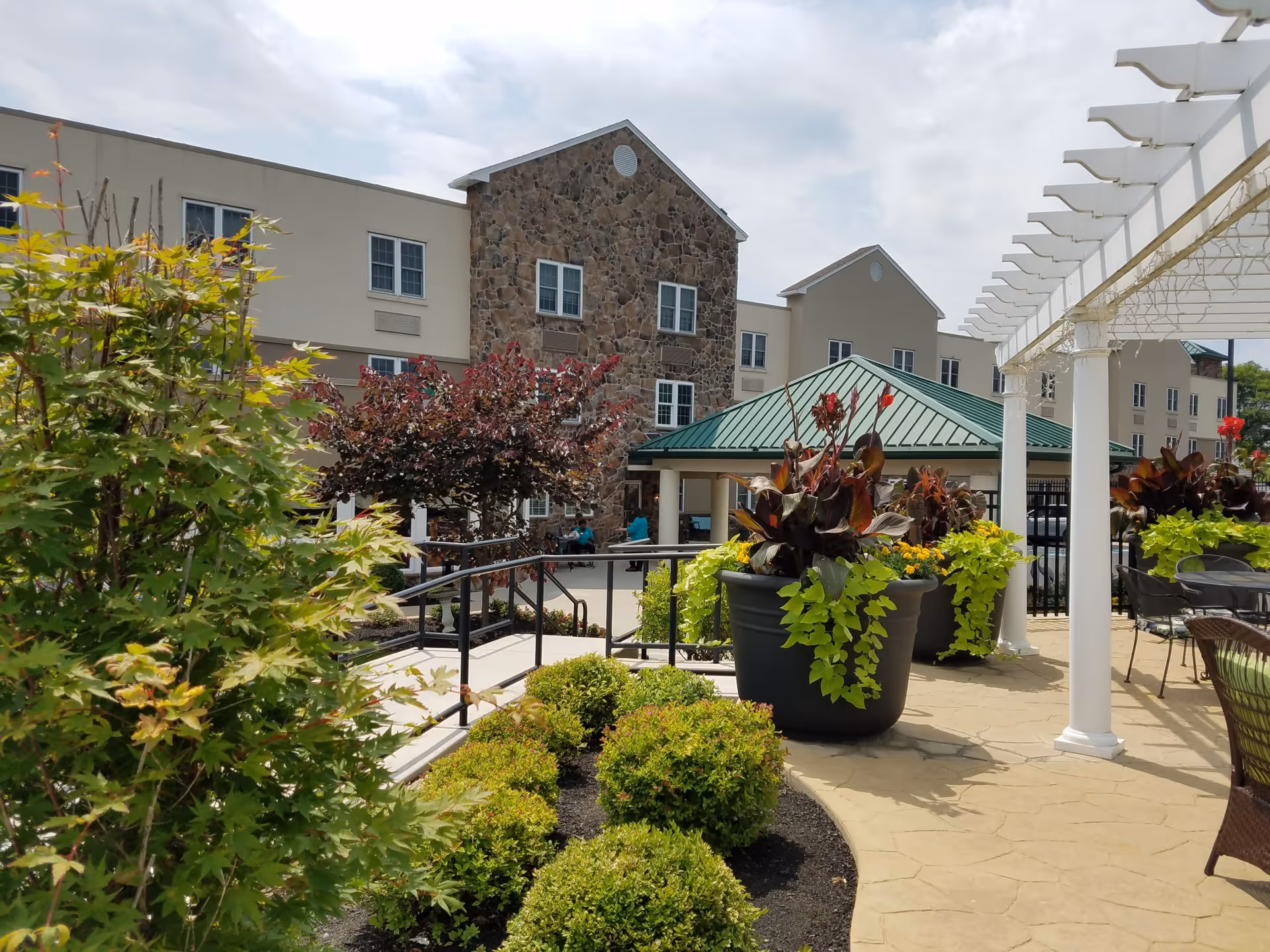 Outdoor patio area at Commonwealth Senior Living At Willow Grove featuring large potted plants, a white pergola, outdoor seating with tables and chairs, and a multi-story building with stone and beige exterior in the background under a partly cloudy sky.
