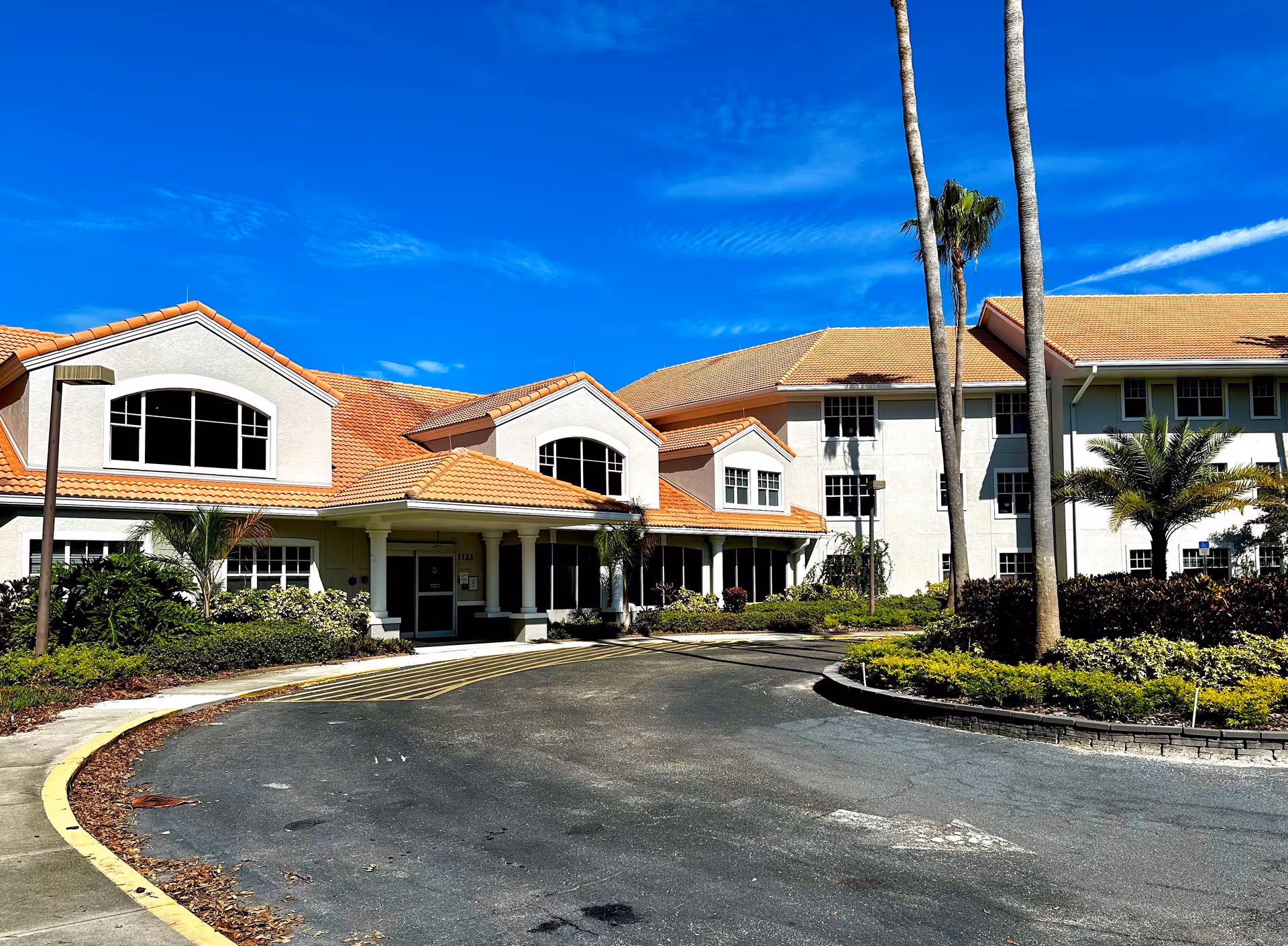 Exterior view of Venice Palms Senior Living facility showing a large building with a tiled roof, multiple windows, palm trees, and landscaped greenery under a clear blue sky.