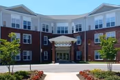Front exterior view of a three-story senior living facility building with a brick facade and white upper walls, featuring a covered entrance with columns, windows on all floors, small trees, and landscaped flower beds along the driveway.