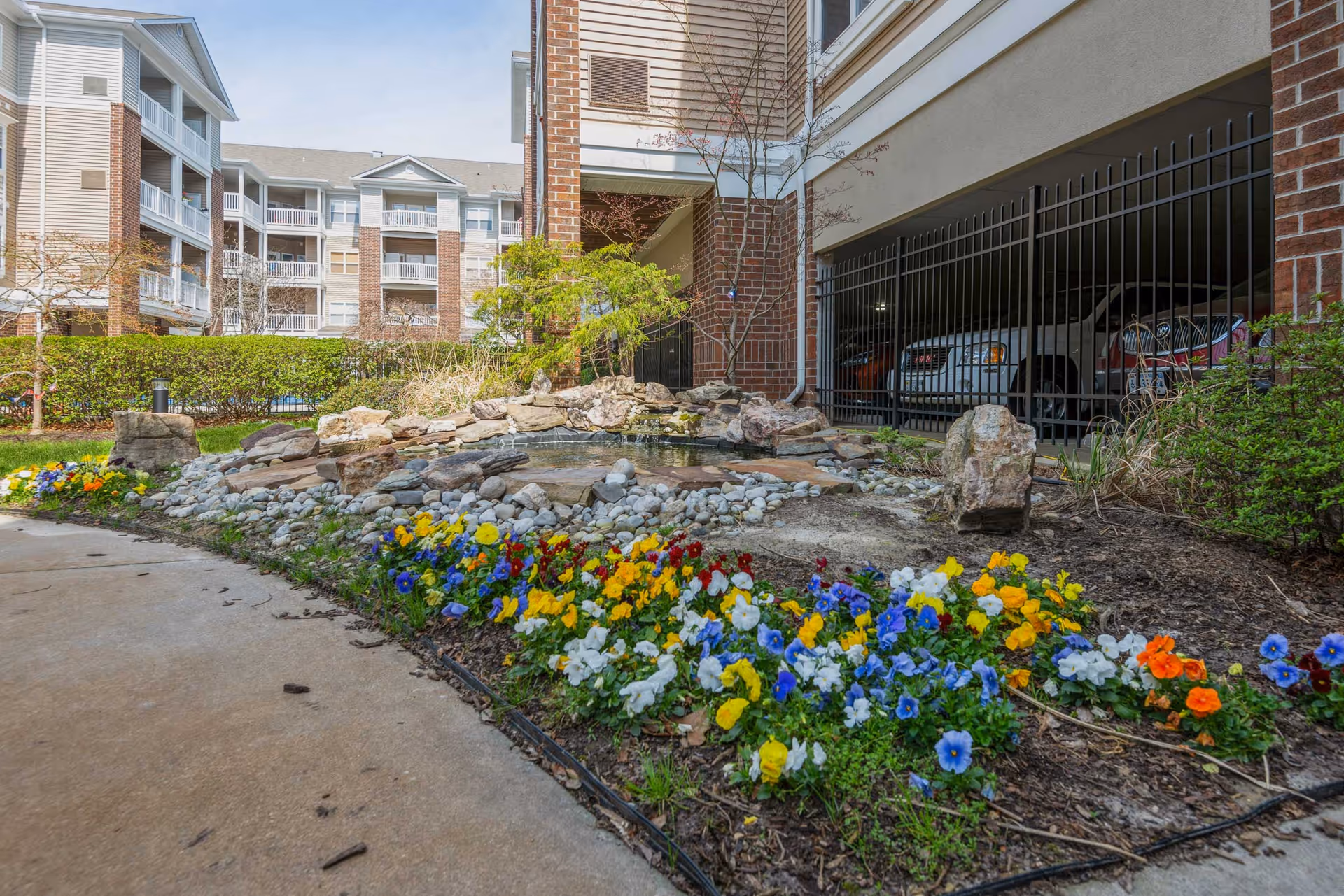 Outdoor garden area at The Talbot on Granby featuring a small rock water feature surrounded by colorful flowers including yellow, blue, white, and orange blooms. In the background, there is a multi-story residential building with balconies and a gated parking area with cars visible.