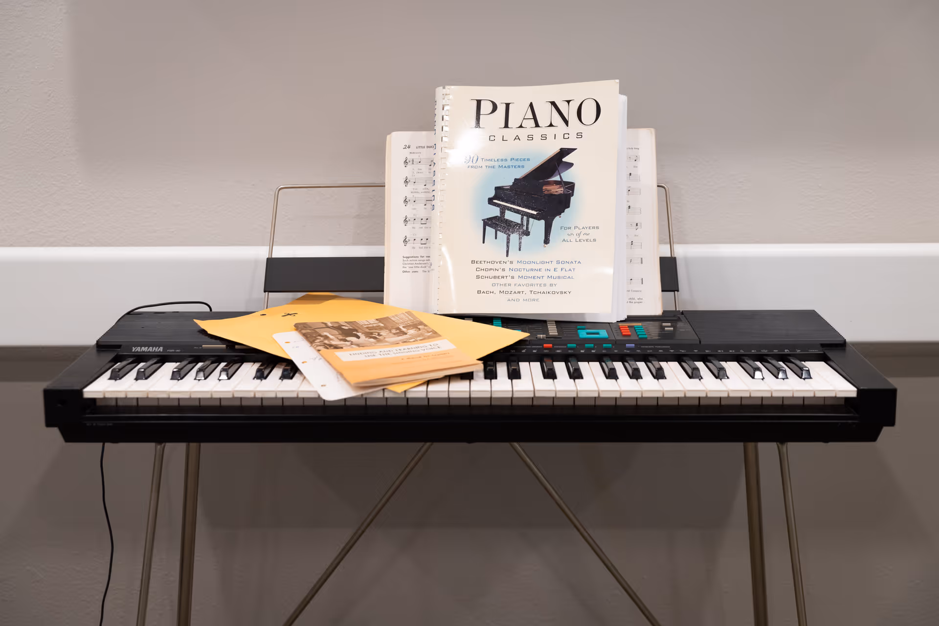 An electronic keyboard on a stand with sheet music and a 'Piano Classics' book propped against a gray wall.