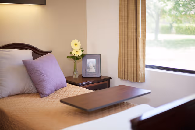 A neatly made bed with a beige quilt and two pillows, one beige and one purple, next to a wooden nightstand holding a small vase with yellow flowers and a framed black and white photo. A window with beige curtains lets in natural light.
