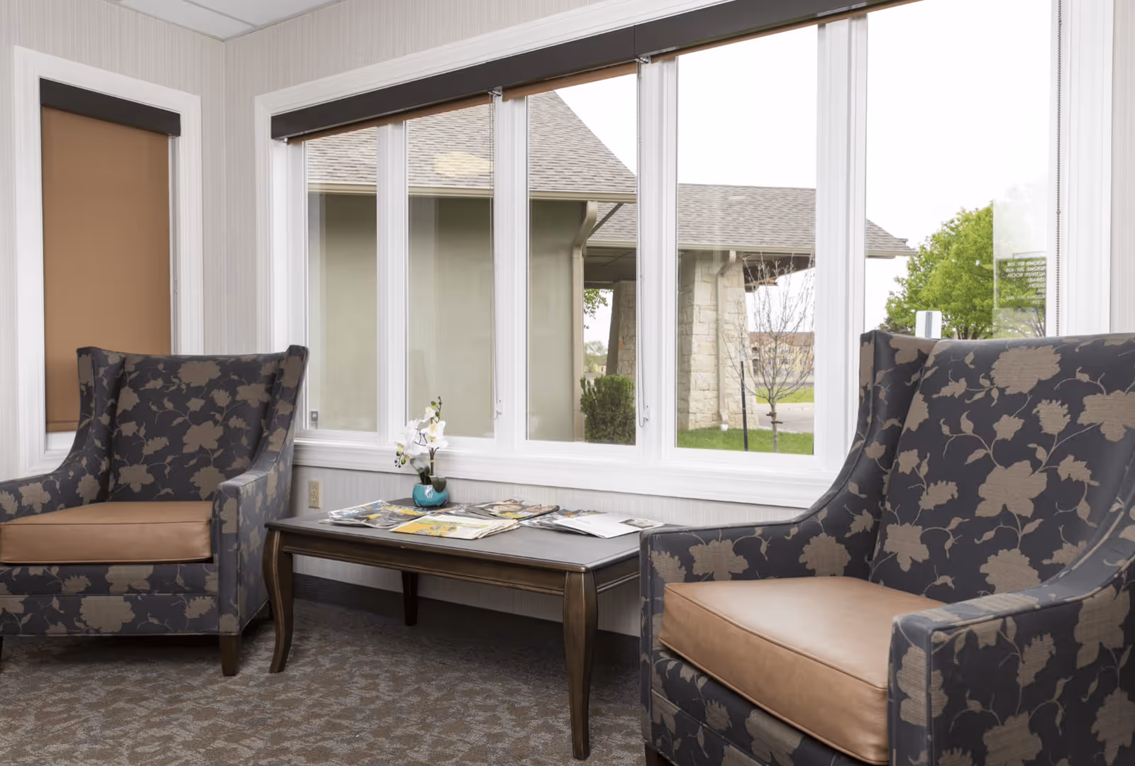 Two patterned armchairs flanking a coffee table with magazines in a bright sitting area by large windows.