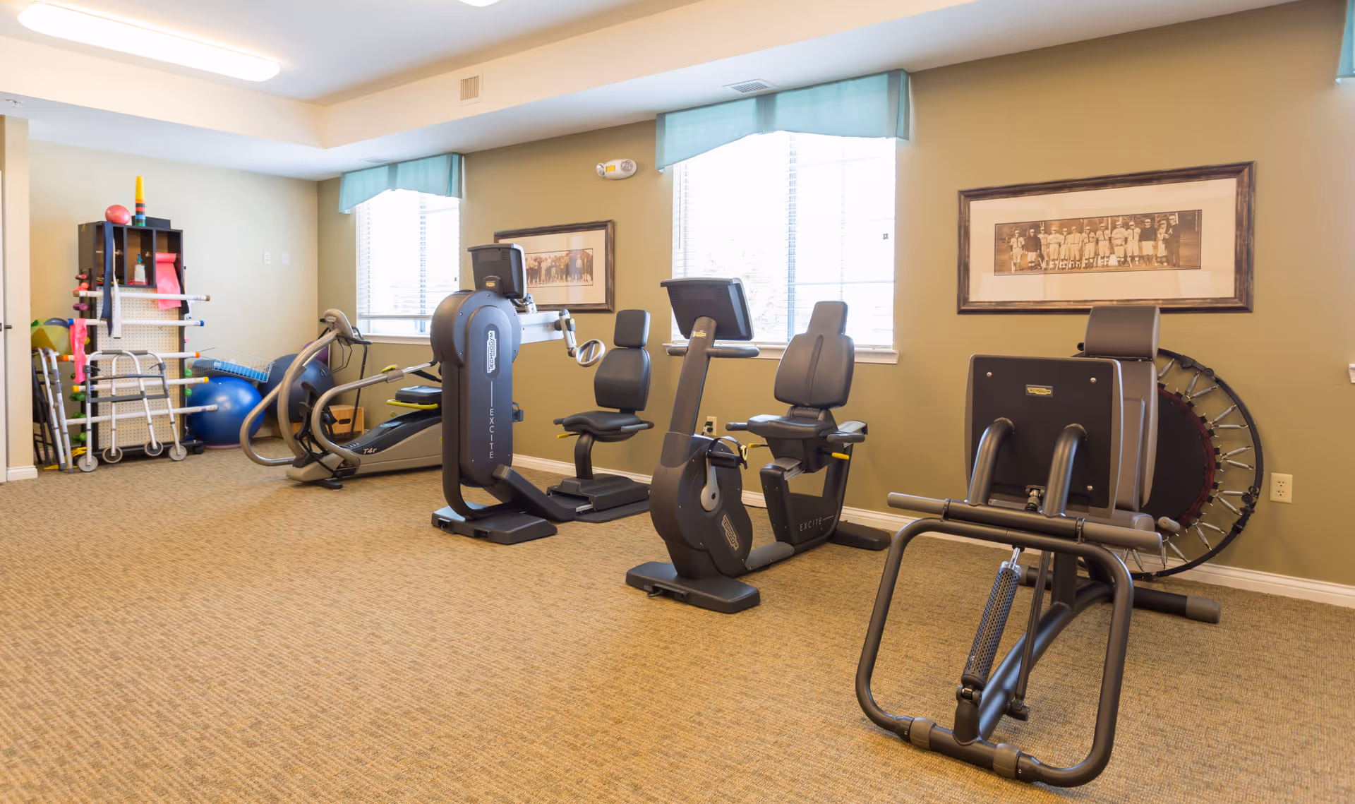A senior living facility exercise room with various fitness equipment including stationary bikes, a rowing machine, and a leg press machine. The room has beige walls, carpeted floor, two windows with light blue valances, framed pictures on the walls, and a shelf with exercise accessories and equipment.