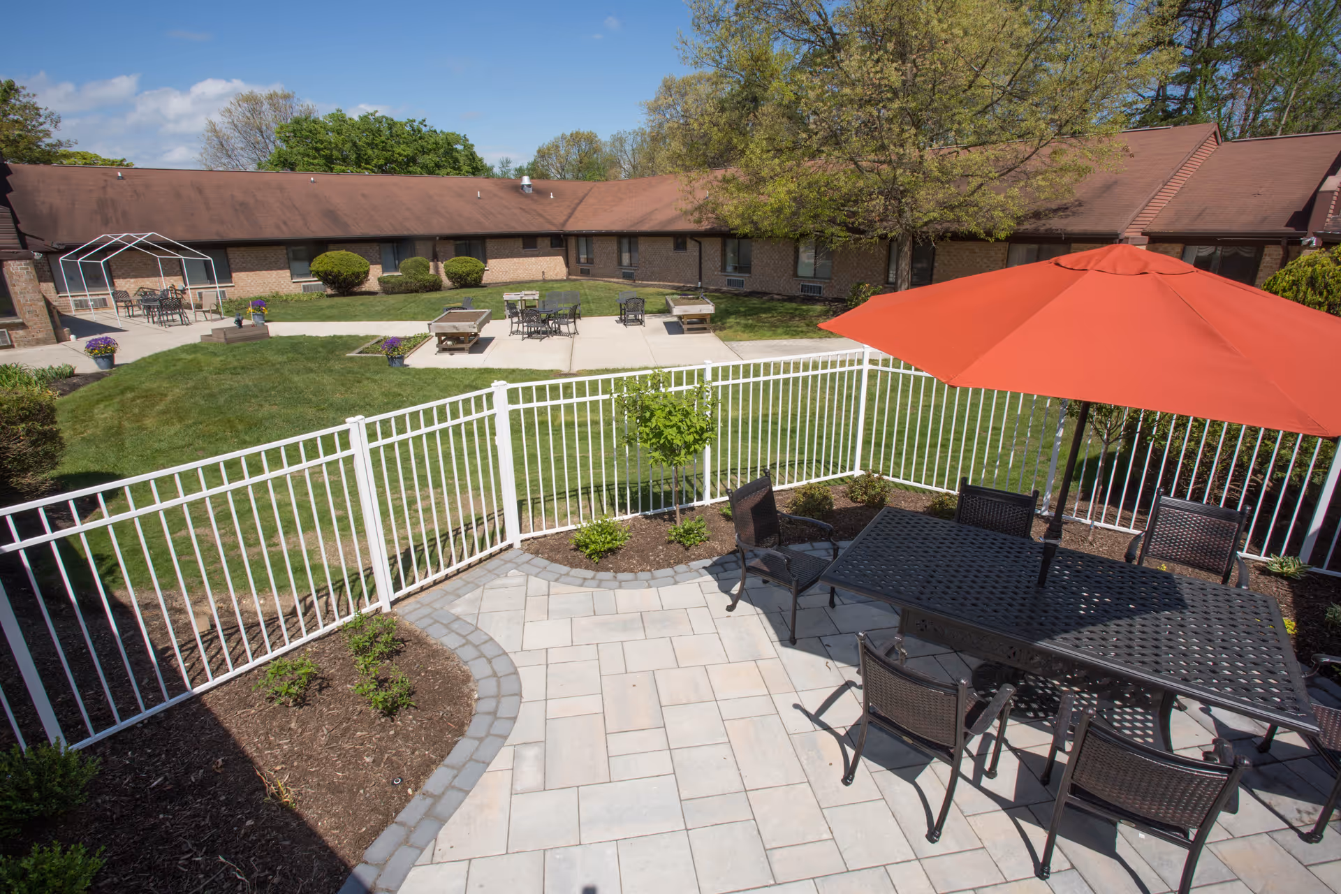 Outdoor patio area with a black metal table and six chairs under a large red umbrella, surrounded by a white fence. Beyond the fence is a grassy courtyard with additional seating areas, small bushes, and a single tree. A one-story brick building with a brown roof encloses the courtyard under a blue sky with some clouds.