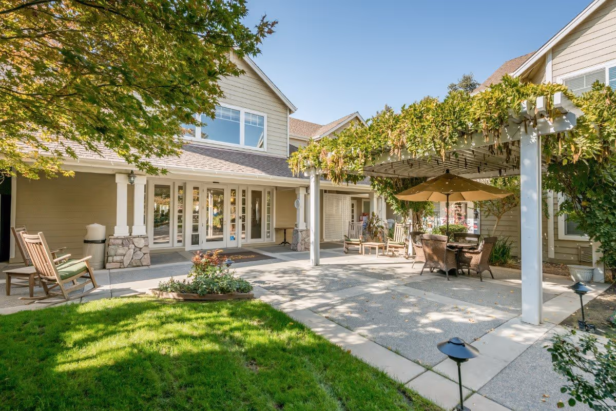 Outdoor patio area at The Grove Senior Living featuring a pergola covered with greenery, a table with an umbrella and chairs, rocking chairs, and a well-maintained lawn with trees and shrubs under a clear blue sky.
