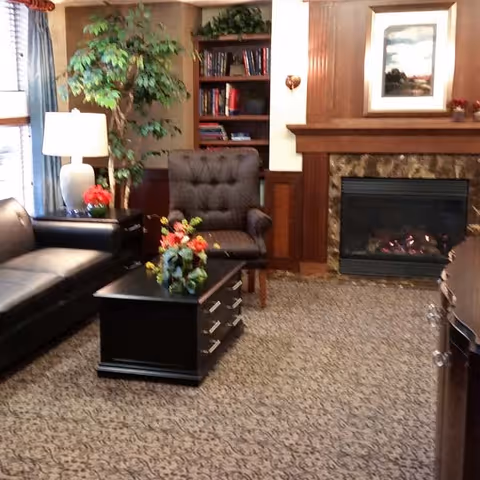 A cozy living room area with a black leather sofa, a dark wooden coffee table with floral arrangements, a tufted armchair, a tall green plant, a table lamp, a built-in bookshelf with books, and a fireplace with a framed picture above it.