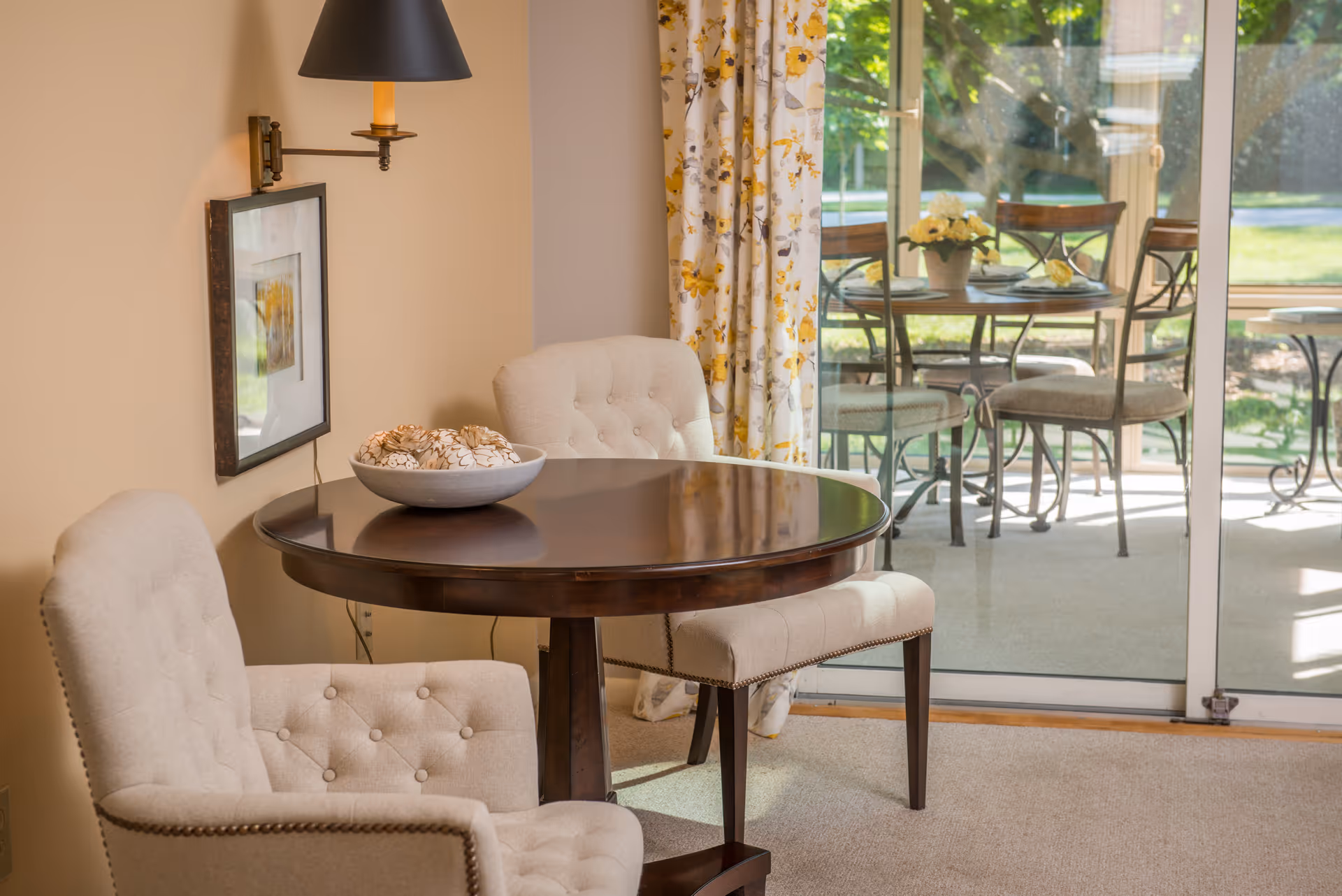 A cozy interior seating area with a round wooden table and two beige tufted chairs. A bowl with decorative items sits on the table. Behind the table, there is a large sliding glass door with floral curtains, leading to an outdoor patio with a metal table and chairs. Sunlight brightens the room.