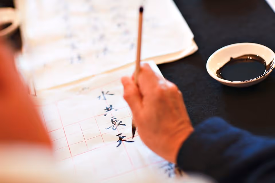 A person practicing calligraphy with a brush on paper that has a grid pattern, with a small dish of black ink placed nearby on a dark surface.
