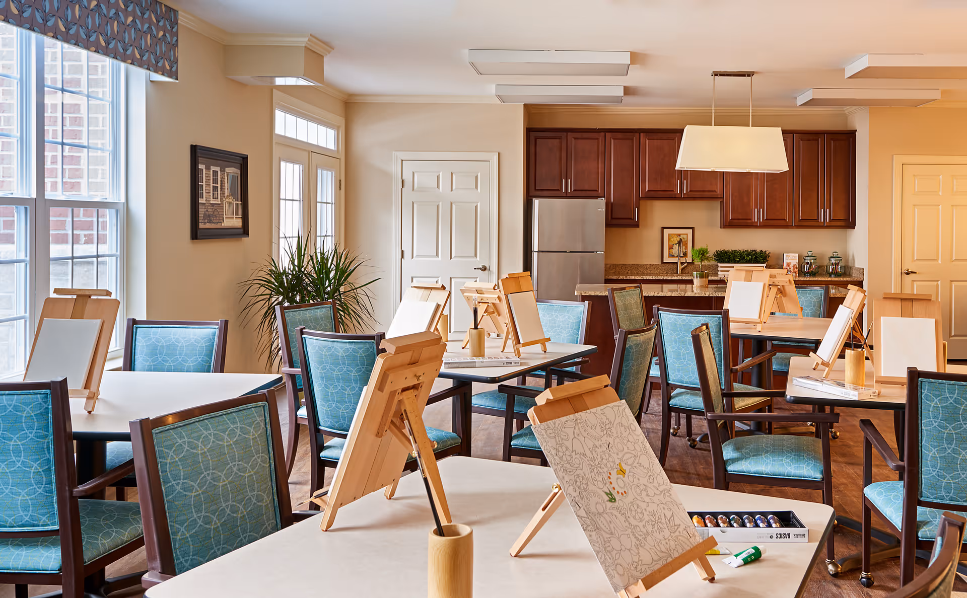 A bright and spacious activity room with multiple tables and chairs arranged for painting. Each table has wooden easels with blank or partially painted canvases, paintbrushes, and paint supplies. The room features large windows letting in natural light, a kitchen area with wooden cabinets and a refrigerator in the background, and a potted plant near the door.