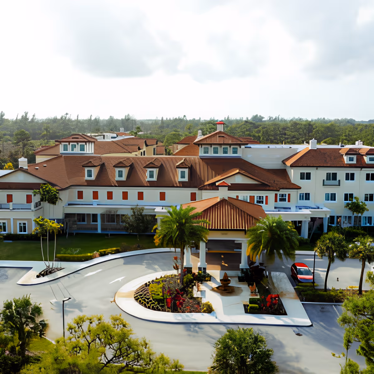 Front view of a multi-story building with red tile roofs, a covered porte-cochere, circular driveway, landscaped island and fountain flanked by palm trees.