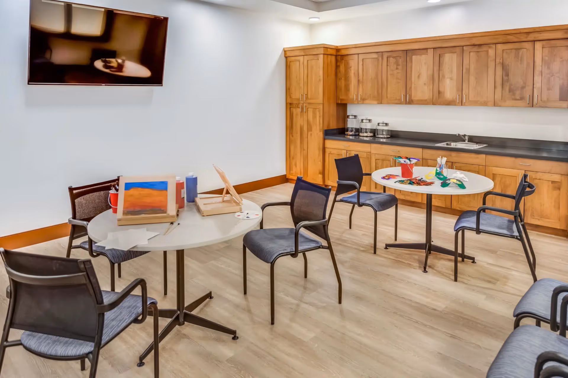 Well-lit interior activity room with round tables and chairs, art supplies and small paintings on the tables, wooden cabinets and a wall-mounted TV.