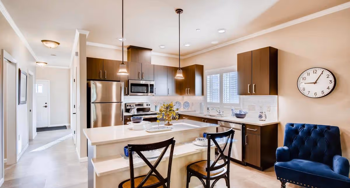 A modern kitchen with dark wood cabinets, stainless steel refrigerator, microwave, and stove. There is a white countertop island with two black chairs, pendant lights hanging above, and a blue upholstered armchair to the right. A round wall clock shows the time as 1:10. The kitchen has a window with white blinds and beige walls.