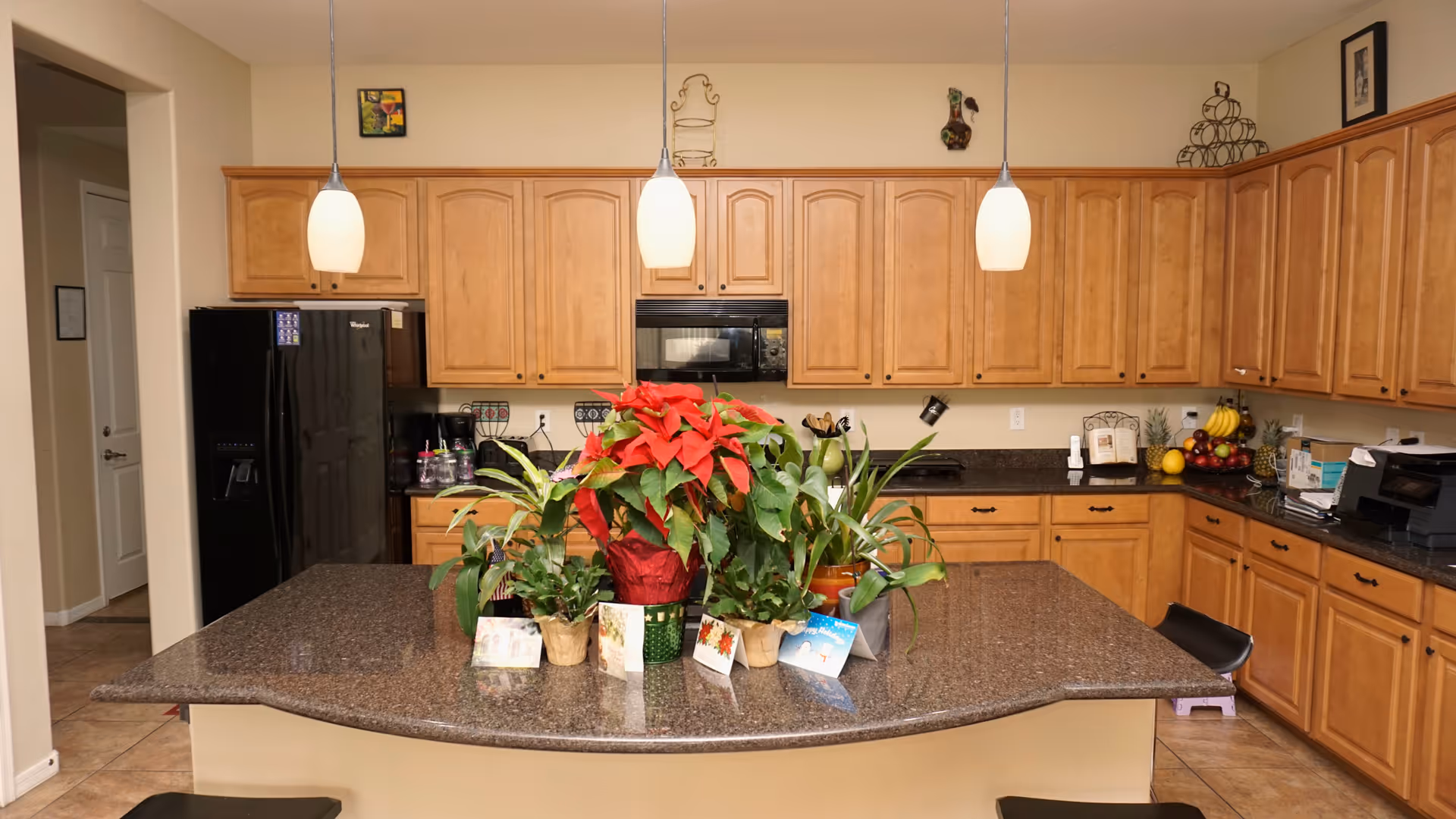 A spacious kitchen with wooden cabinets and a large granite countertop island in the center. The island is decorated with several potted plants and greeting cards. The kitchen features a black refrigerator, a built-in microwave, and various kitchen items on the countertops. Three pendant lights hang above the island, and the floor is tiled.