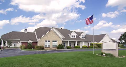 Single-story senior living facility building with a large lawn, an American flag on a pole, and a partly cloudy sky.