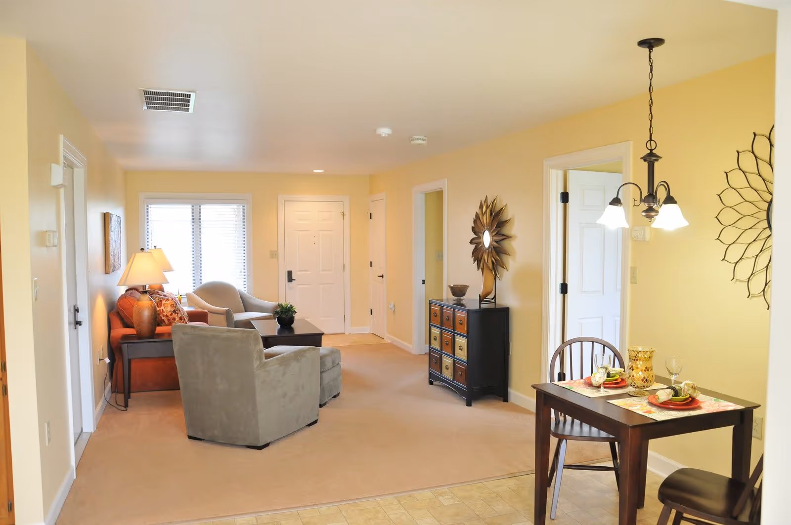 Open senior living and dining area with armchairs and a sofa, a small table set for two, decorative wall accents, and an entry door.