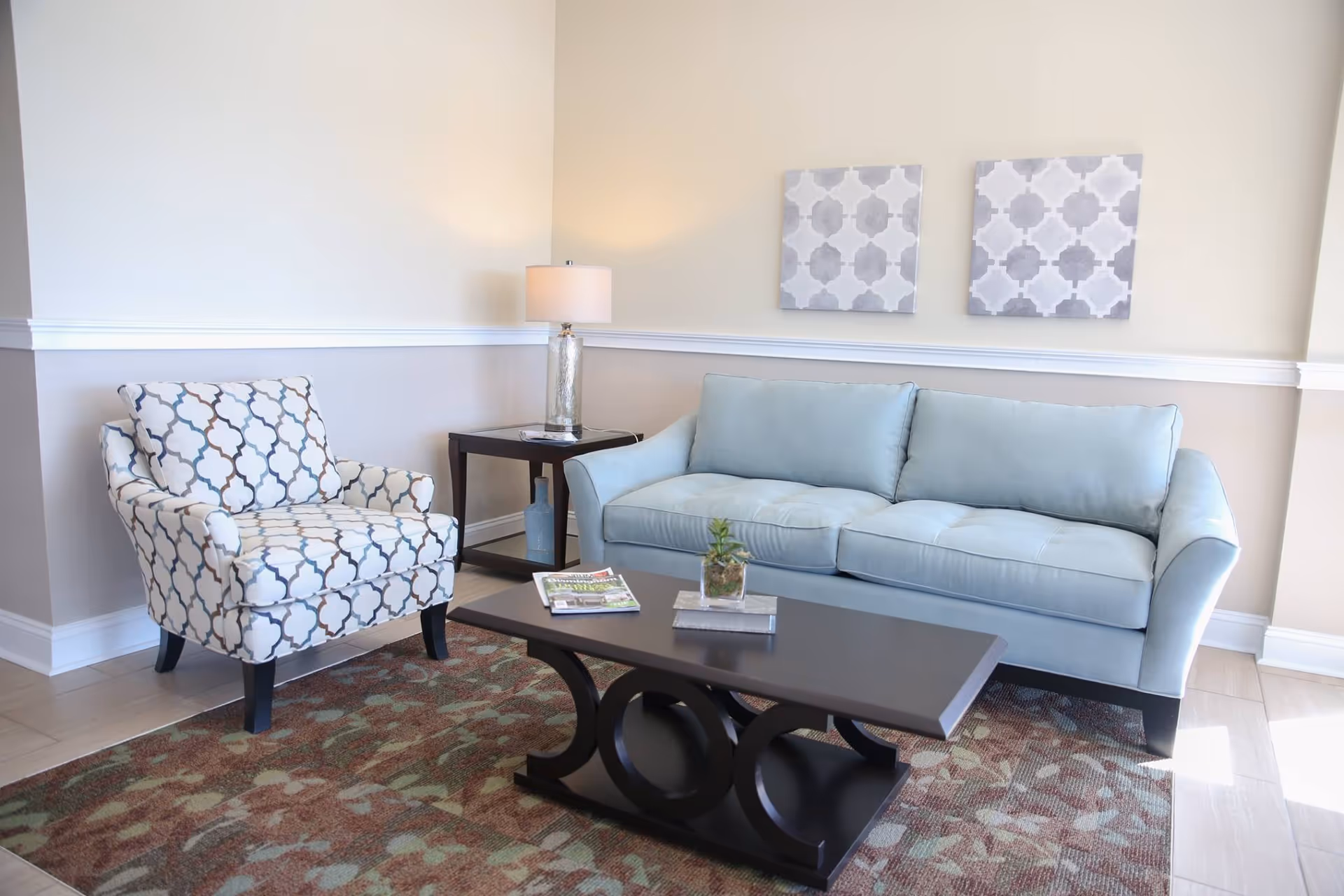 A cozy living room area with a light blue sofa, a patterned armchair, a dark wooden coffee table with a small plant and magazines on it, a side table with a lamp, and two decorative wall art pieces above the sofa.