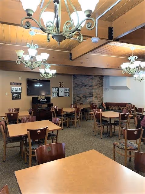 Interior view of a senior living facility common area with multiple wooden tables and chairs arranged neatly. The room features several decorative chandeliers hanging from a wooden beam ceiling, a brick fireplace in the corner, a flat-screen TV mounted on the wall, and framed pictures surrounding the TV. There is also a striped couch near the fireplace.
