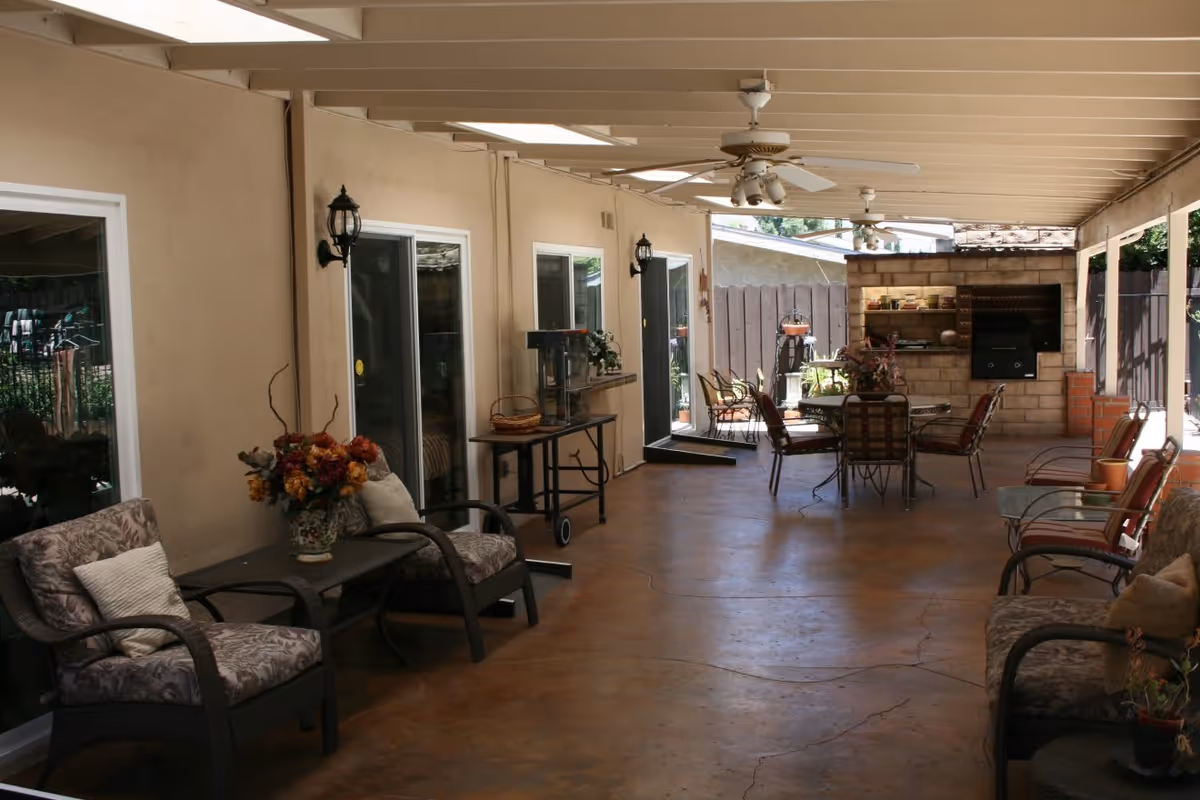 Covered outdoor patio area with several cushioned chairs and small tables arranged along the sides. Ceiling fans are mounted on the white ceiling beams. At the far end, there is a built-in brick barbecue grill and a round table with chairs. The patio floor is a smooth concrete surface, and the area is adjacent to a beige building with sliding glass doors and wall-mounted lantern-style lights.