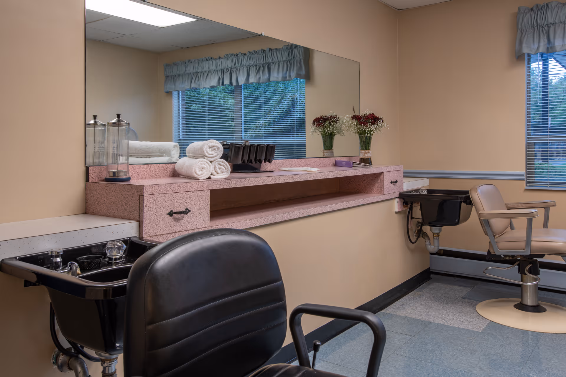 Interior view of a hair salon area in a senior living facility with two salon chairs, black shampoo sinks, a large mirror, rolled white towels, and vases with flowers on a pink countertop. There are windows with blinds and beige walls.