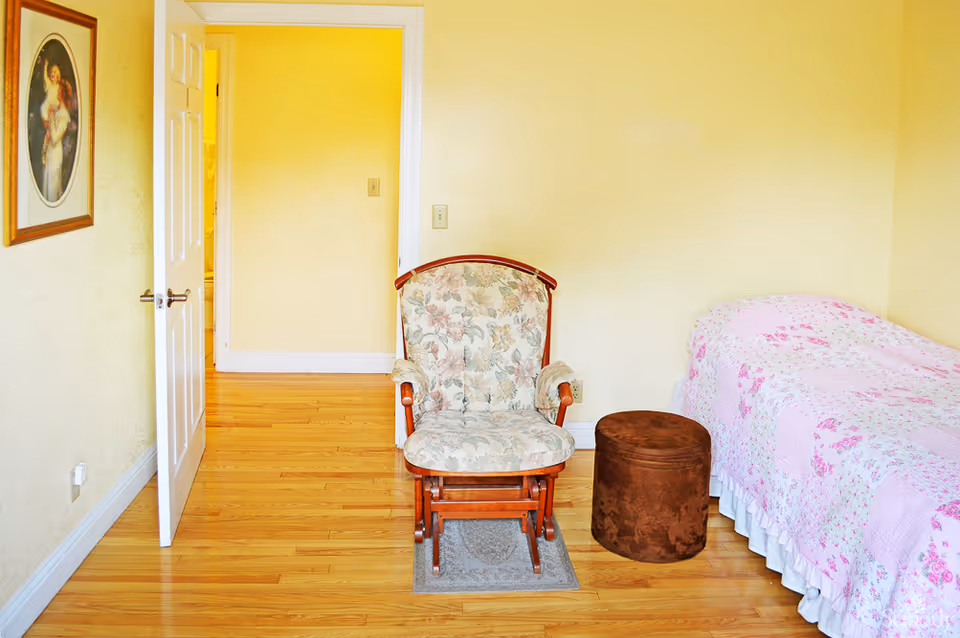 A bright bedroom with a single floral quilted bed, a patterned upholstered chair on a small rug, and a brown ottoman on hardwood floors.