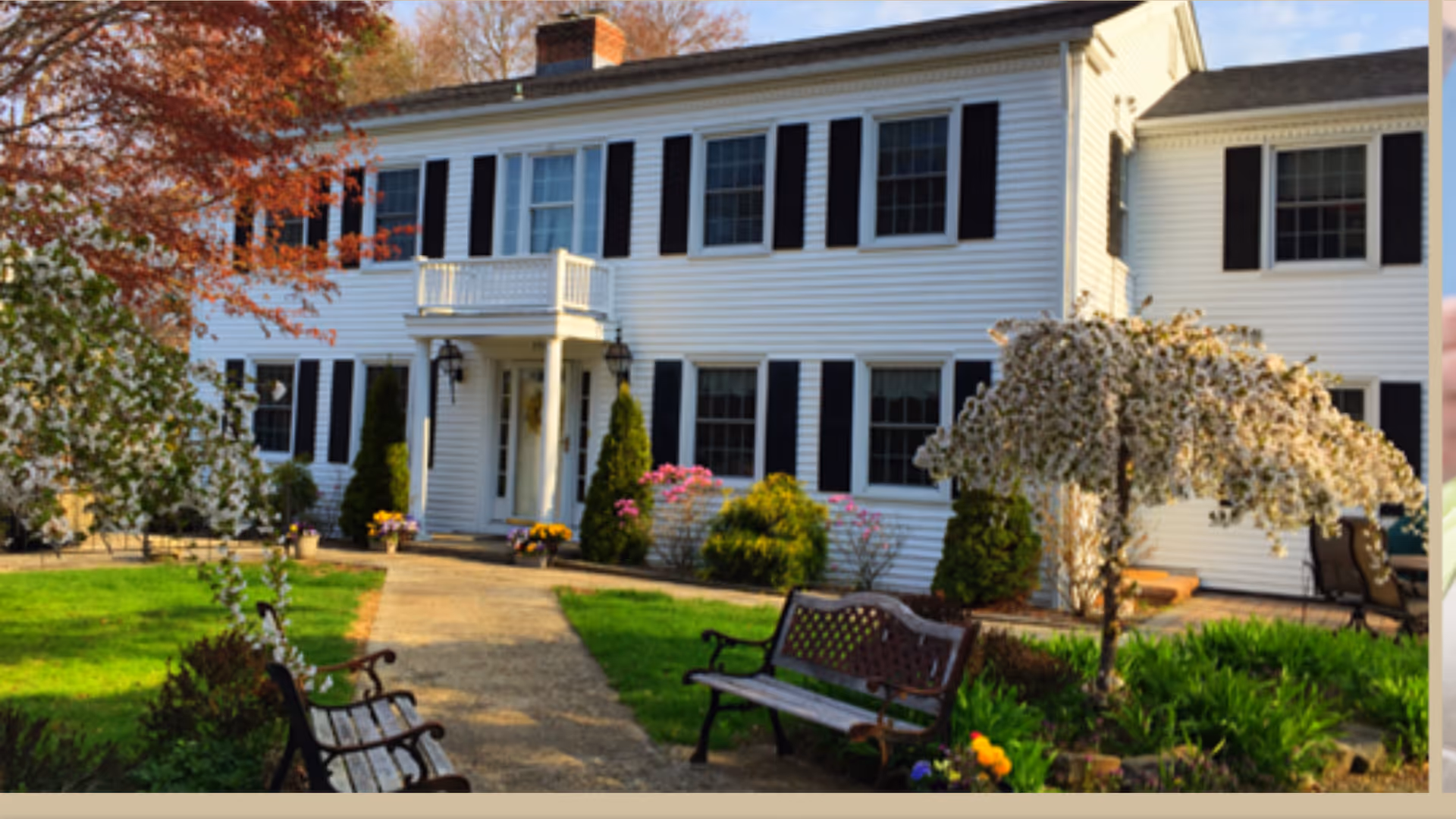 Front exterior view of a white two-story building with black shutters, a small balcony above the entrance, and a well-maintained garden with blooming trees, shrubs, flowers, and benches along a paved walkway.