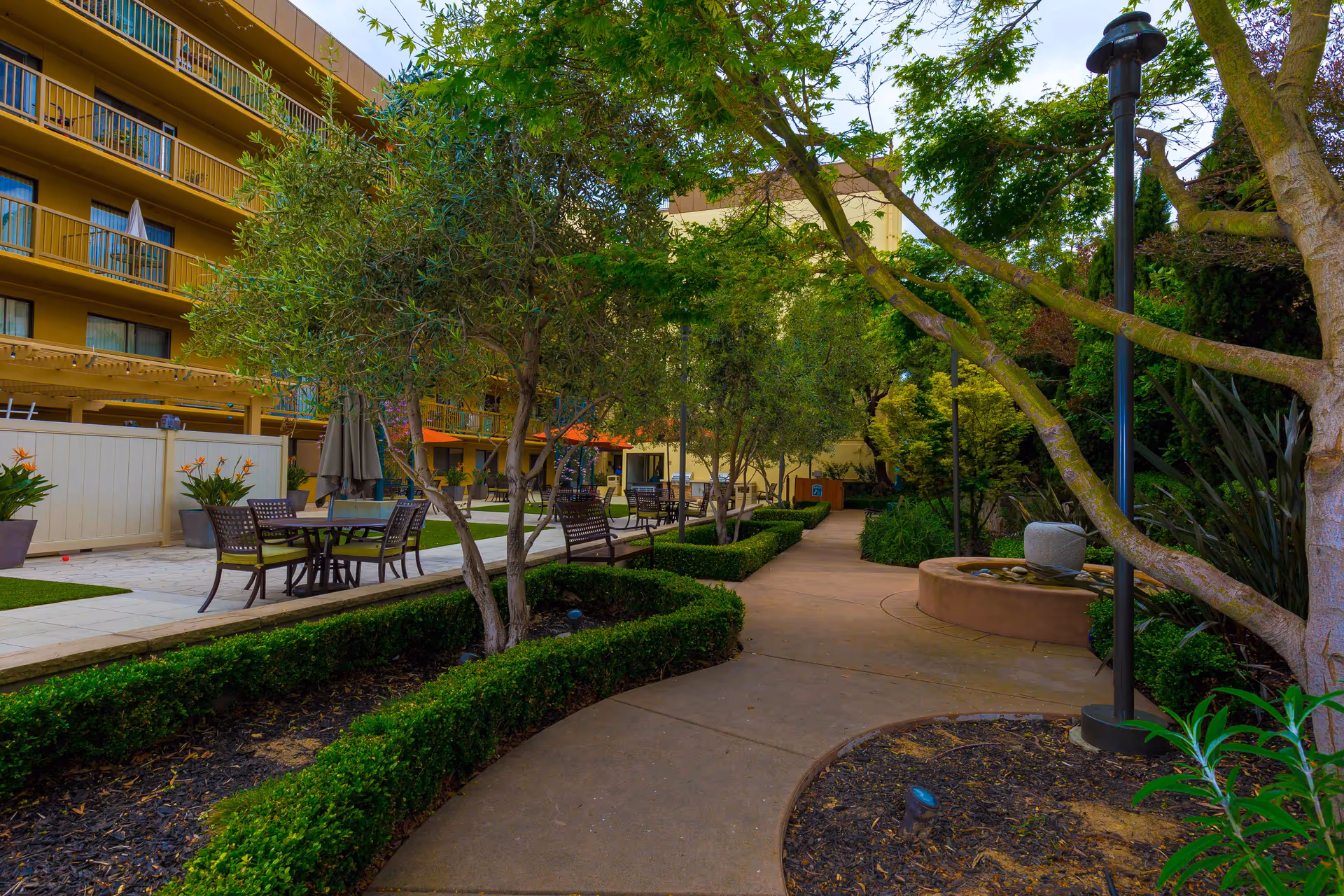 Outdoor garden area at The Point At Rockridge Senior Living featuring a paved walkway, green hedges, trees, benches, tables with chairs, and a small water fountain under a partly cloudy sky.