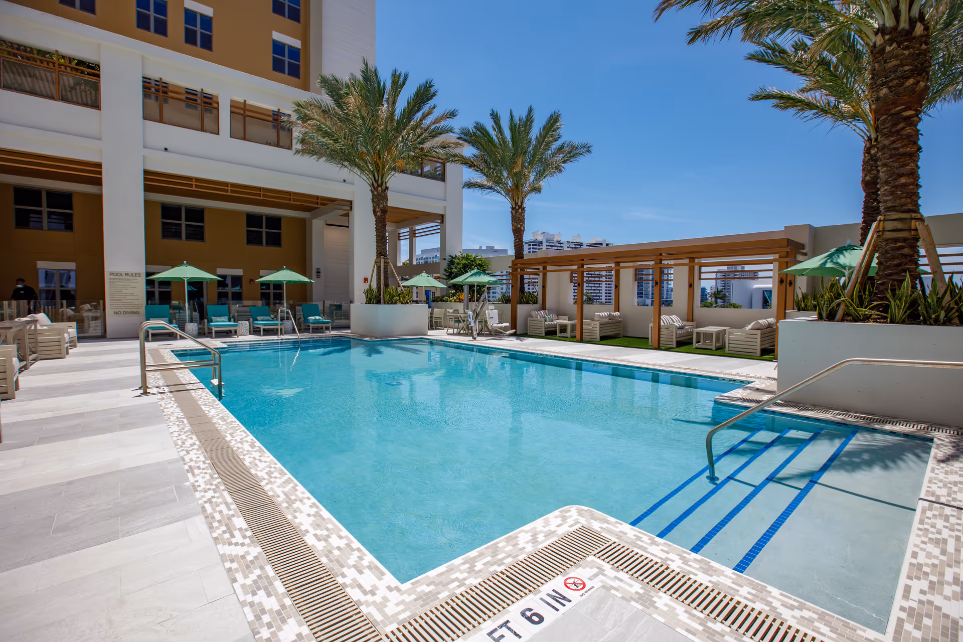 Outdoor swimming pool area with lounge chairs, green umbrellas, palm trees and cabanas next to a multi-story building.