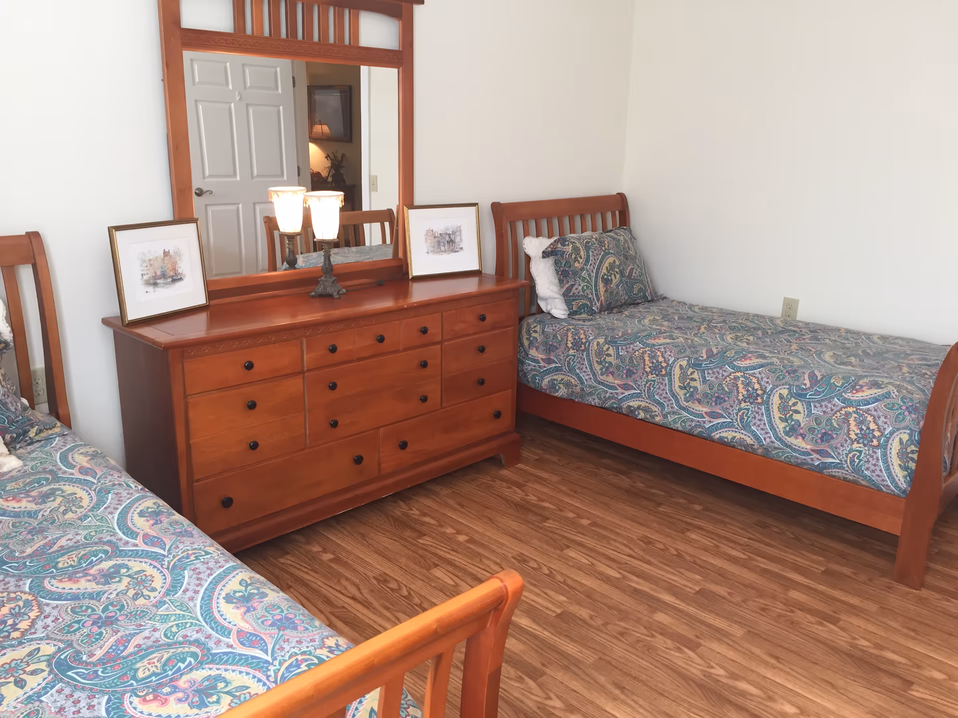 Two twin beds with paisley bedspreads and a wooden dresser with mirror and lamp in a bright bedroom.
