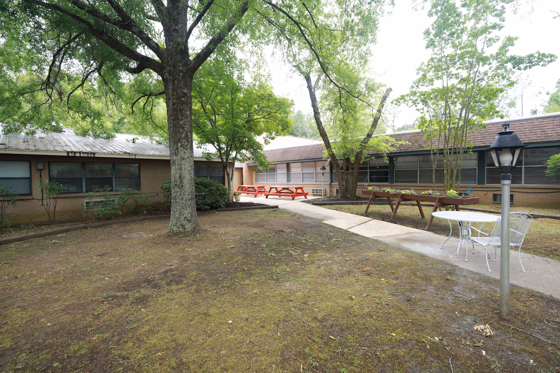 Outdoor courtyard area at Spring Gate Rehabilitation and Health Care Center featuring a large tree in the center, a concrete walkway, red picnic tables, raised garden beds, a white metal table with chairs, and a lamp post. The courtyard is surrounded by single-story building wings with windows and a brown roof.