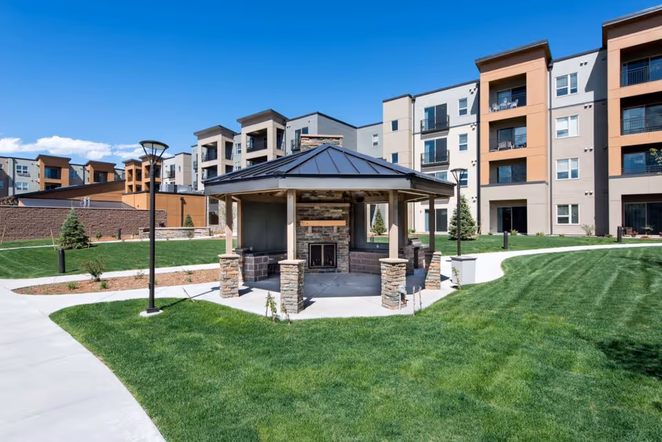 Outdoor view of a senior living facility with a central gazebo featuring a stone fireplace, surrounded by well-maintained green lawns, paved walkways, modern street lamps, and multi-story residential buildings in the background under a clear blue sky.