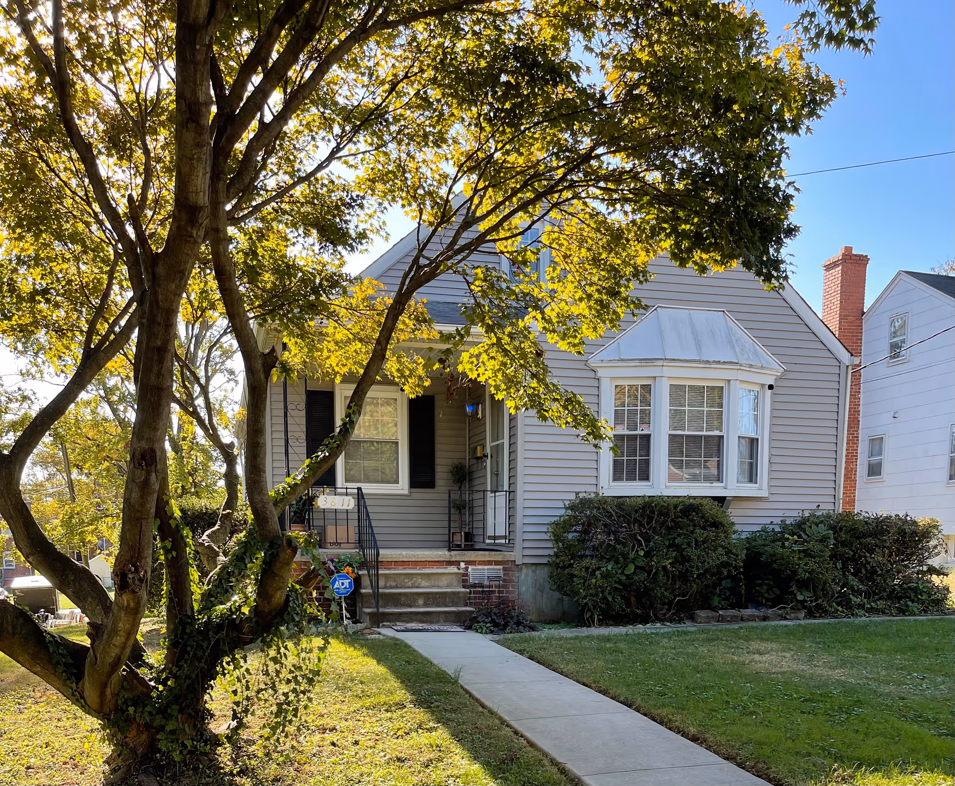 A small residential house with gray siding, a bay window, and a front porch with steps leading up to the door. There is a large tree with green leaves in the front yard, casting shadows on the grass and sidewalk. The house has black shutters and a brick chimney. The sky is clear and blue.