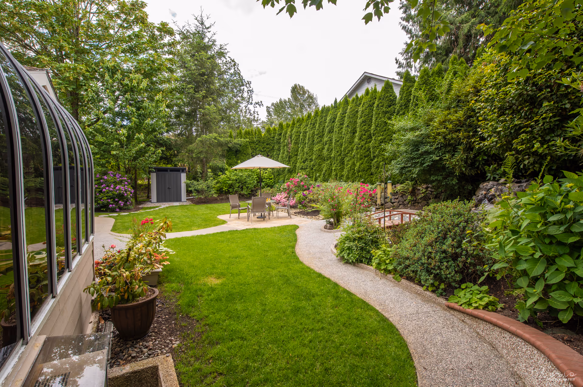 Well-maintained backyard garden with a curved gravel path leading to a patio set with an umbrella, surrounded by lush shrubs and trees.