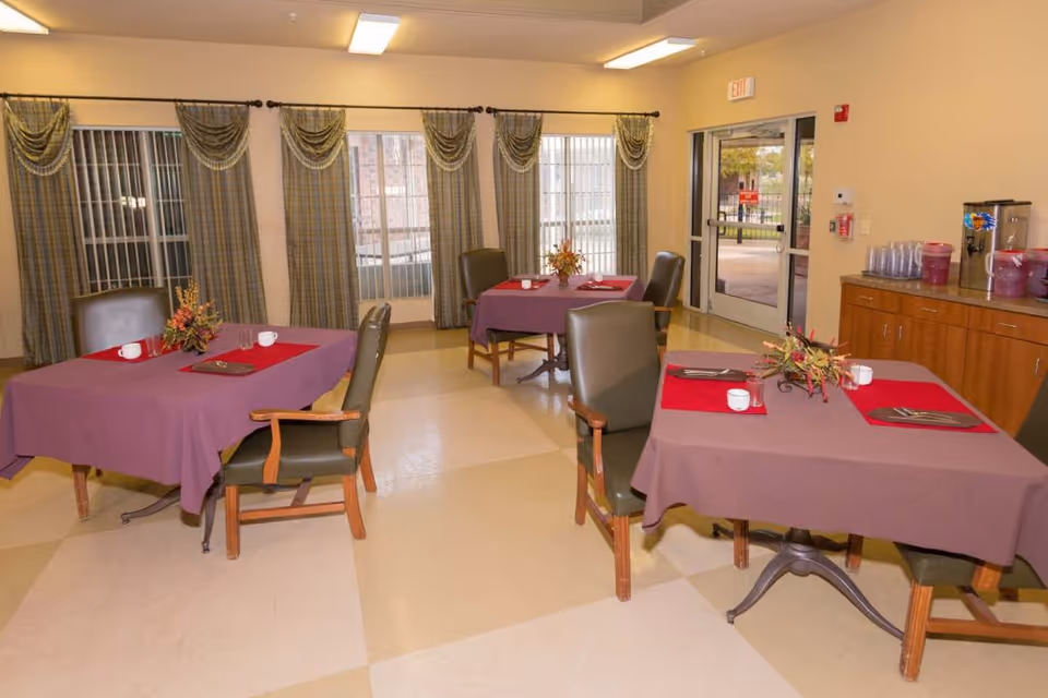 A dining room with three tables covered in purple tablecloths and red placemats, each set with white cups and silverware. The room has large windows with patterned curtains and a door leading outside. There is a wooden cabinet on the right side with beverage dispensers and cups.