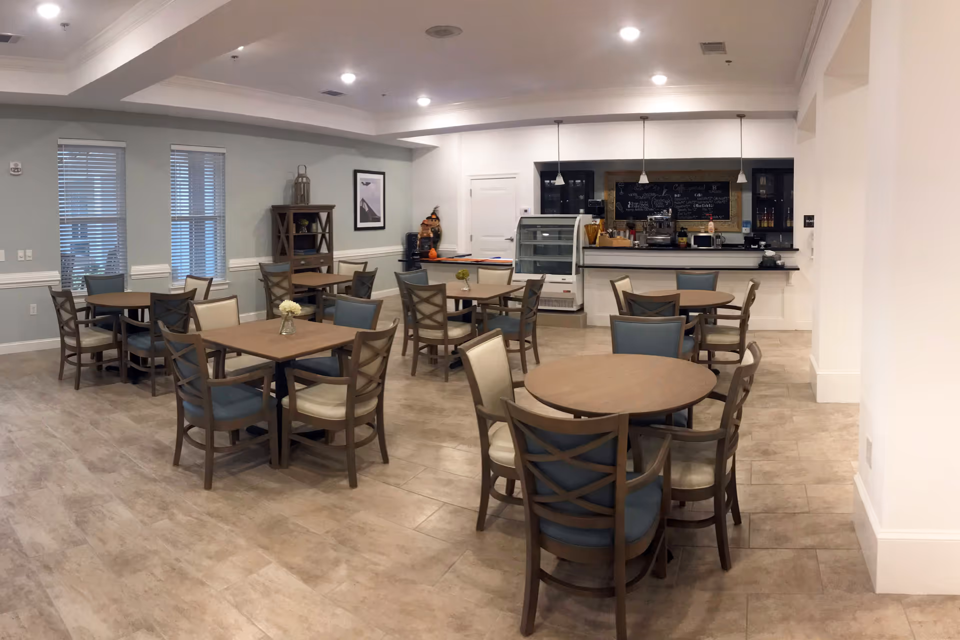 A dining area in a senior living facility with several round and square wooden tables surrounded by chairs with blue and beige cushions. The room has light-colored tile flooring, light blue walls, and recessed ceiling lights. There is a counter area at the back with a display case, coffee machine, and a chalkboard menu. A small shelving unit and framed artwork are visible on the left wall.