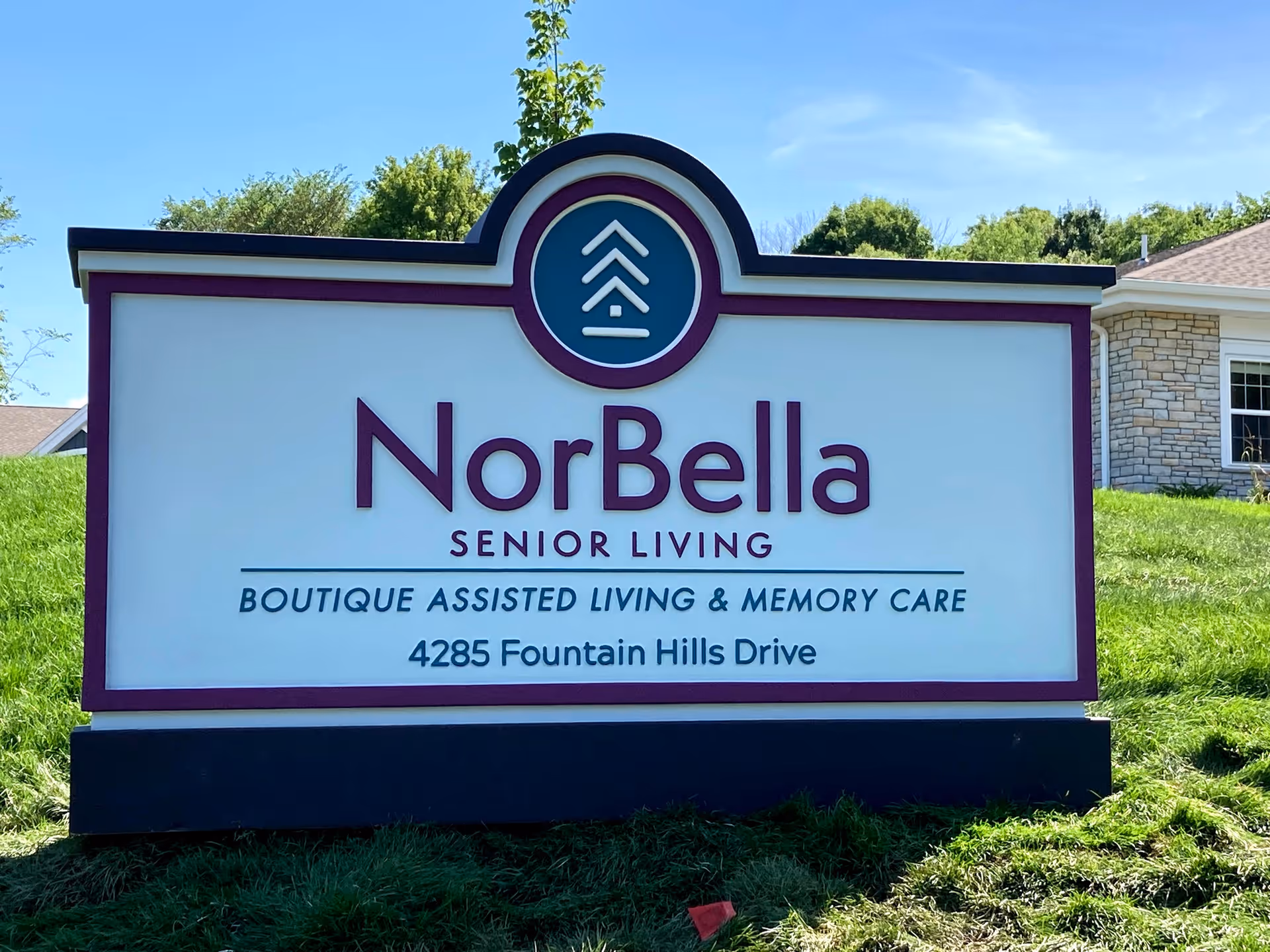 Outdoor sign for NorBella Senior Living, a boutique assisted living and memory care facility located at 4285 Fountain Hills Drive, with a clear blue sky and greenery in the background.