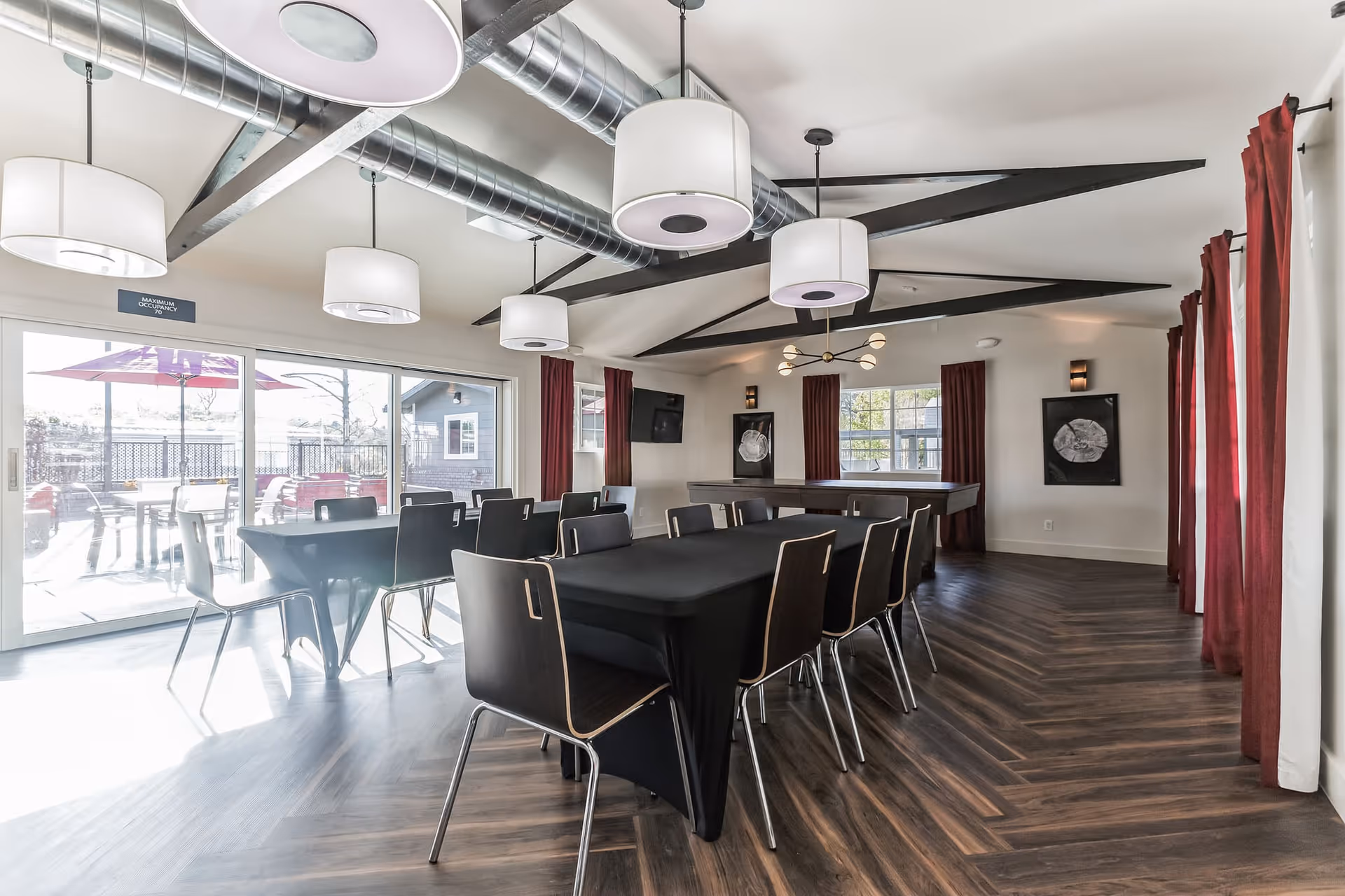 A modern dining room with a long table covered with a black tablecloth surrounded by black chairs. The room features large windows with red curtains, exposed ceiling beams, and multiple pendant lights. Outside the sliding glass doors, there is a patio area with tables and umbrellas.