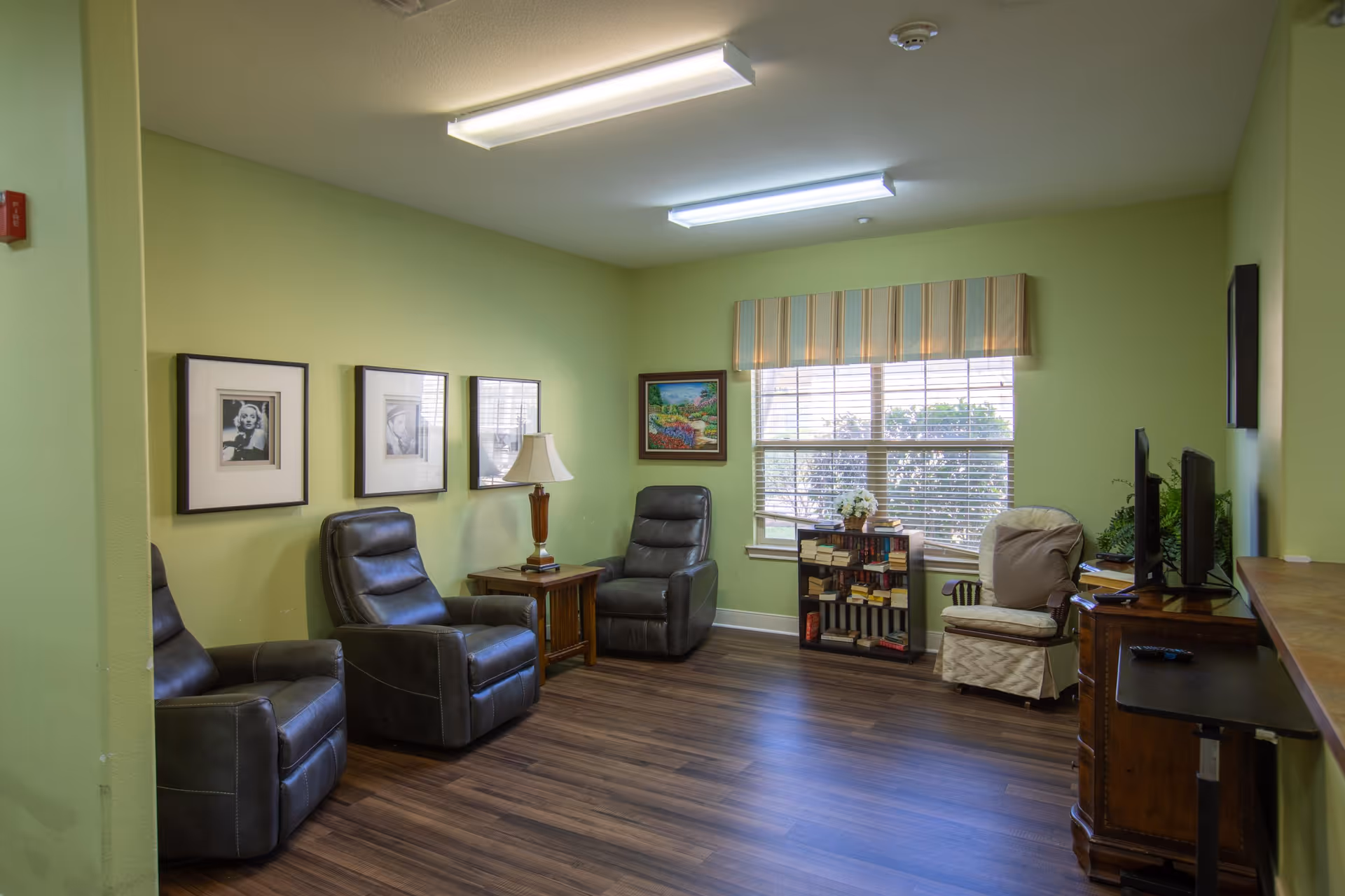 A bright green-walled common room with several recliners, a side table and lamp, a bookcase by a window, and a TV.